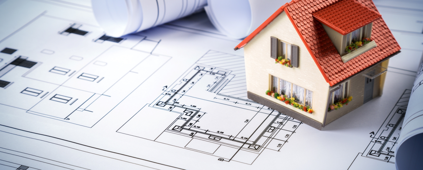Makieta domu jednorodzinnego z czerwonym dachem ustawiona na rozłożonych planach architektonicznych, w tle zwinięte projekty techniczne